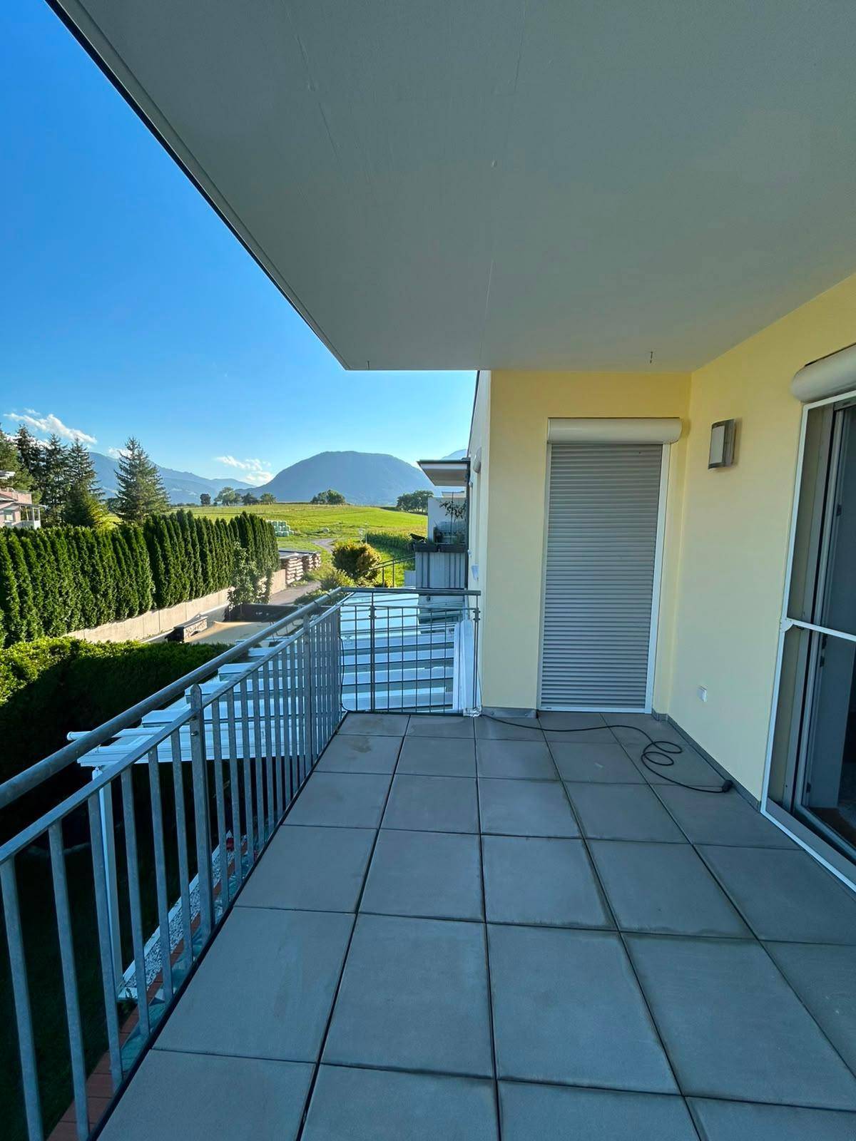 Großer Balkon mit Fliesenboden und weitem Blick über die grüne Landschaft und eine Hecke.