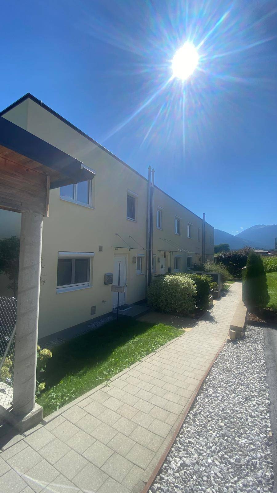 Seitenansicht des Reihenhauses mit gepflastertem Weg und Schotterfläche unter strahlendem Sonnenschein.