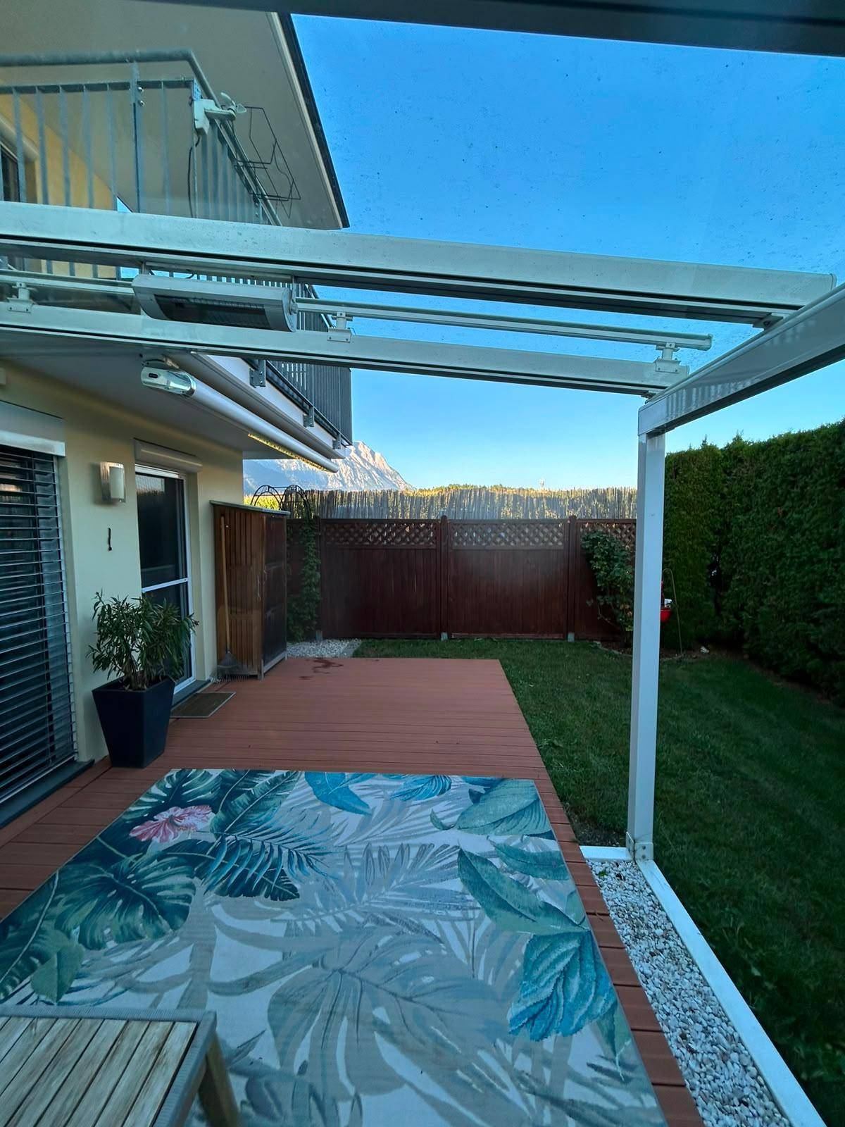 Überdachte Terrasse mit Holzdeck und Gartenblick in der Abenddämmerung, umgeben von hohen Hecken.