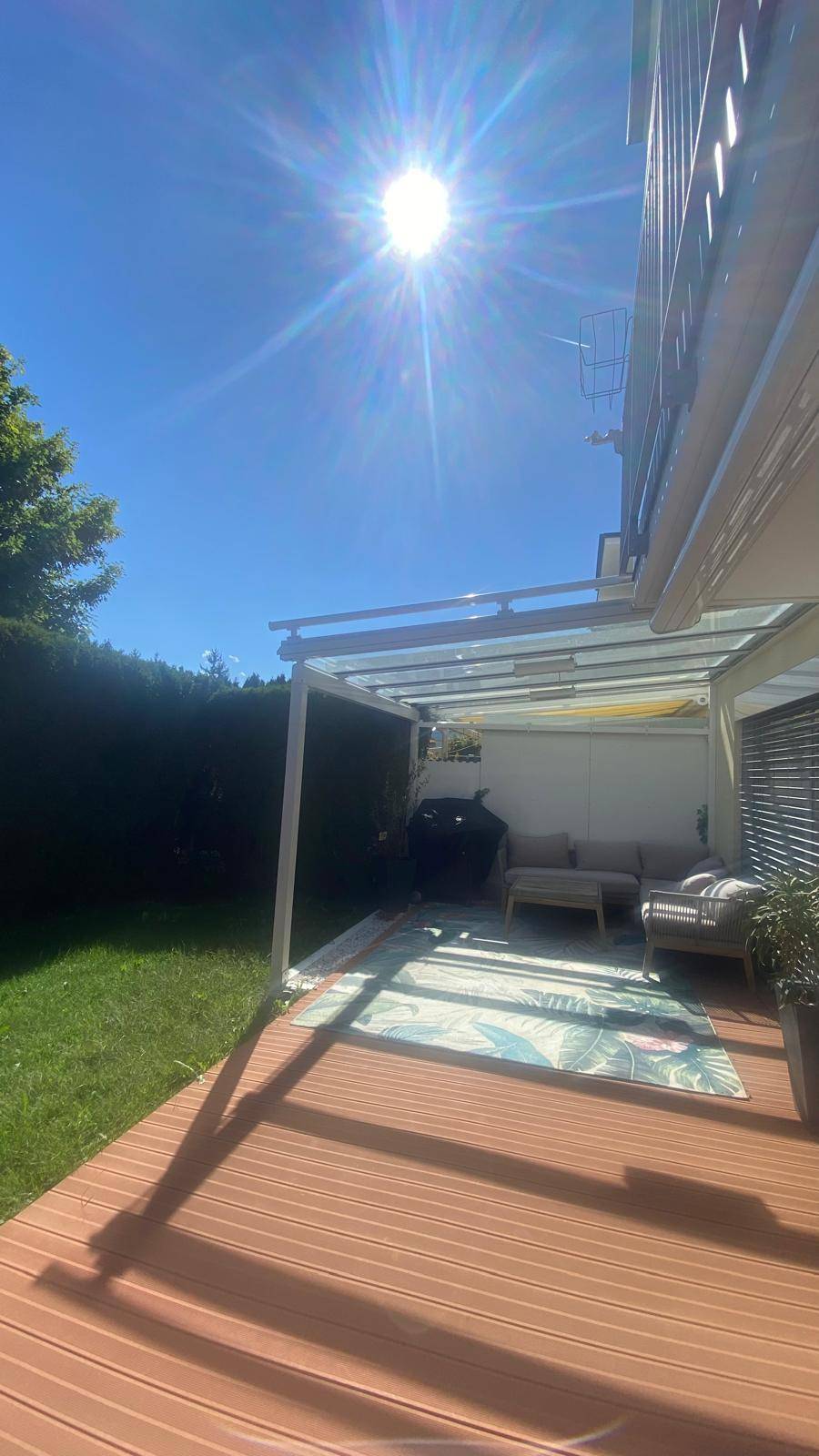 Sonnige Terrasse mit Holzdeck, Gartenmöbeln und einer Überdachung, umgeben von Grünflächen.