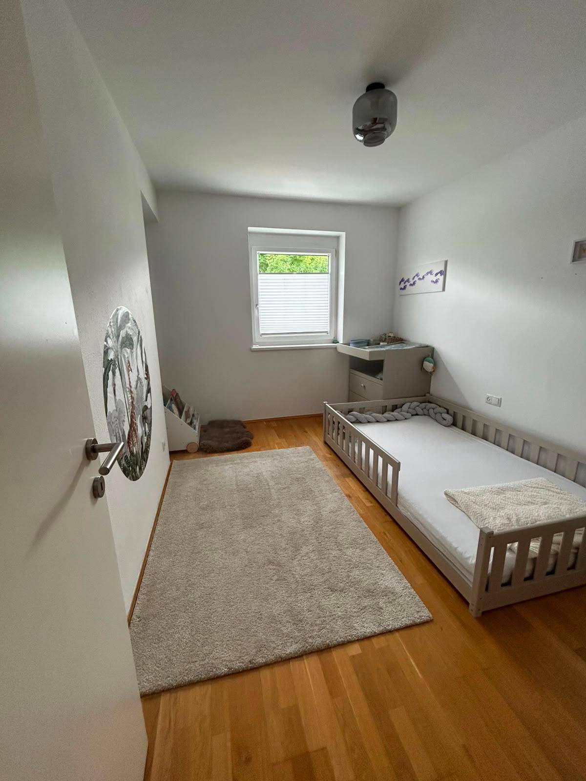 Helles Kinderzimmer mit Holzboden, kleinem Bett und Fenster mit Blick ins Grüne.