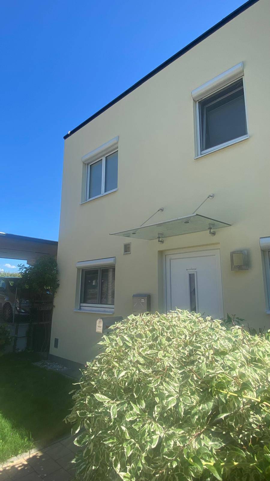 Vorderansicht des Hauses mit Eingangsbereich, Carport und grüner Bepflanzung unter blauem Himmel.