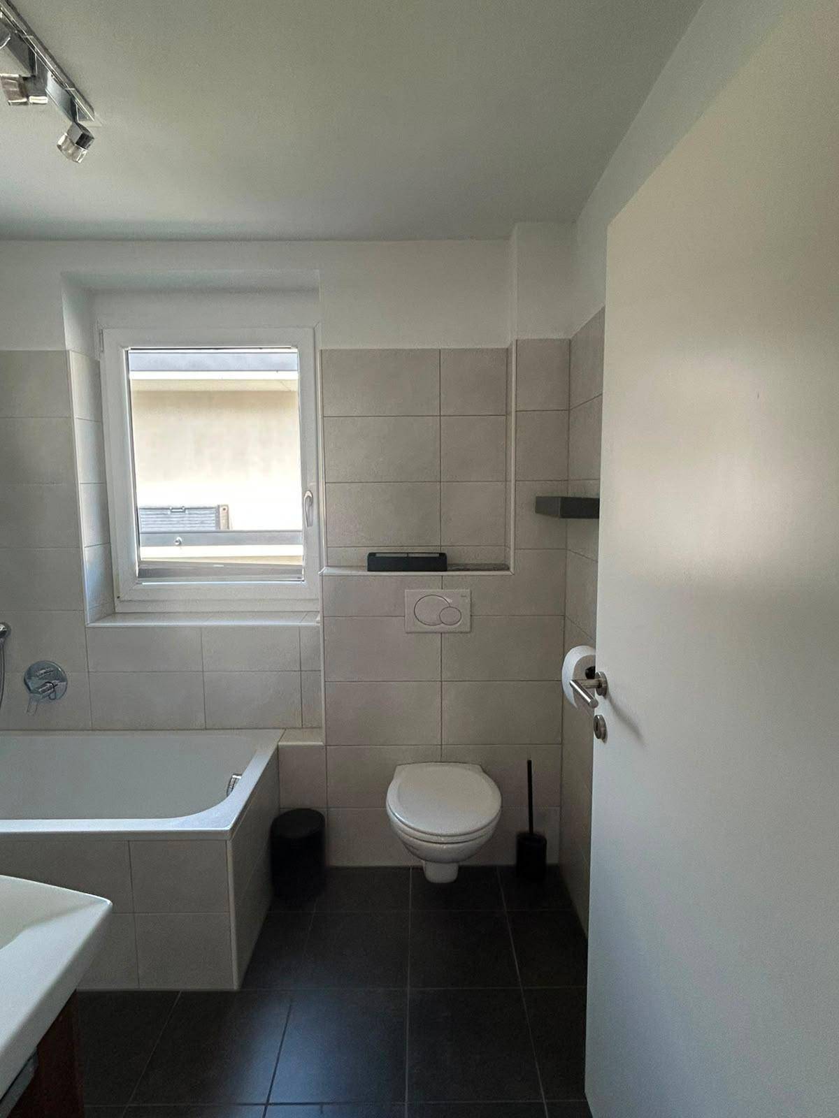 Modernes Badezimmer mit Badewanne, WC, Fenster und dunklen Bodenfliesen.