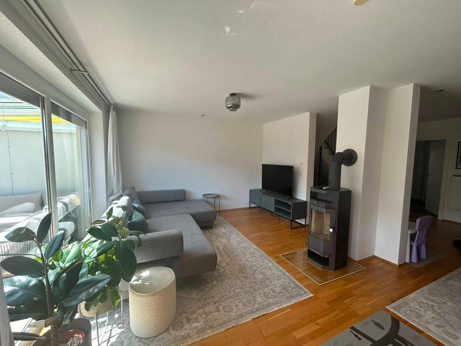 Geräumiges Wohnzimmer mit modernem Sofa, Fernseher und einem gemütlichen Holzofen.