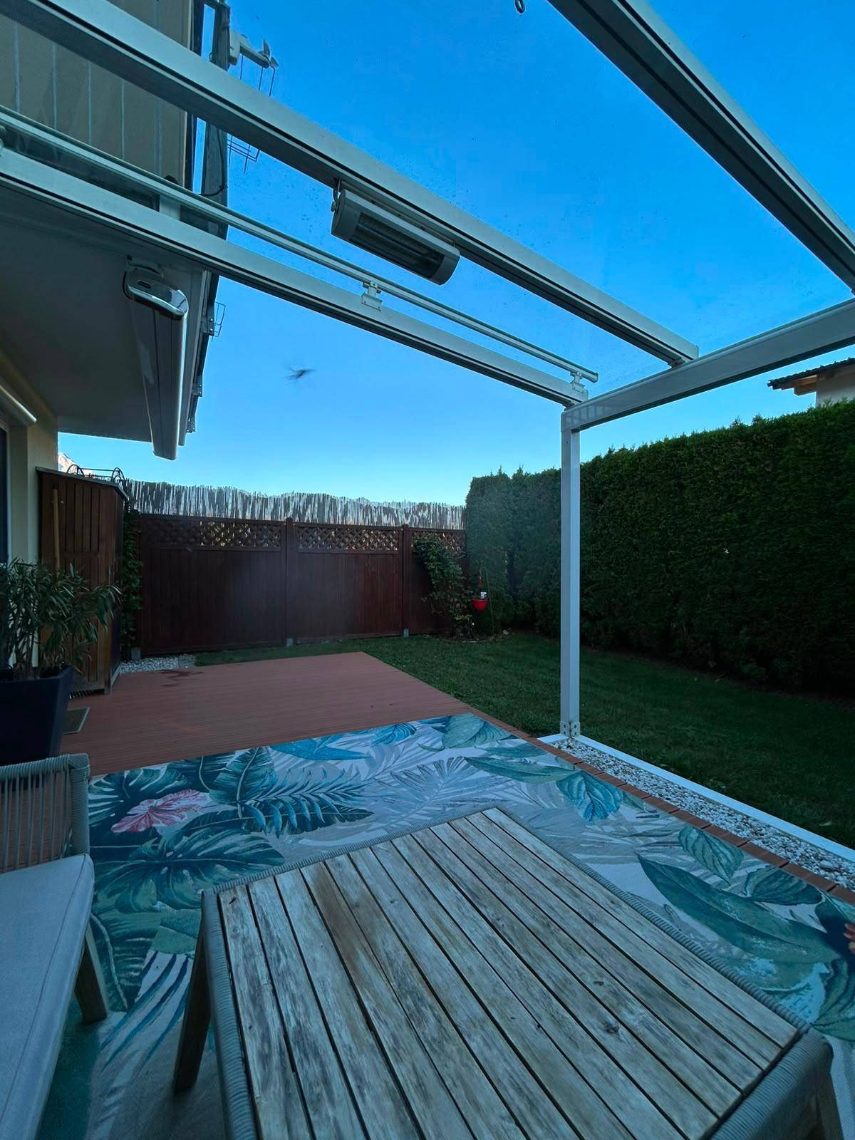 Gemütliche Terrasse mit Holzmöbeln und Teppich, umgeben von einem gepflegten Garten und Hecken.