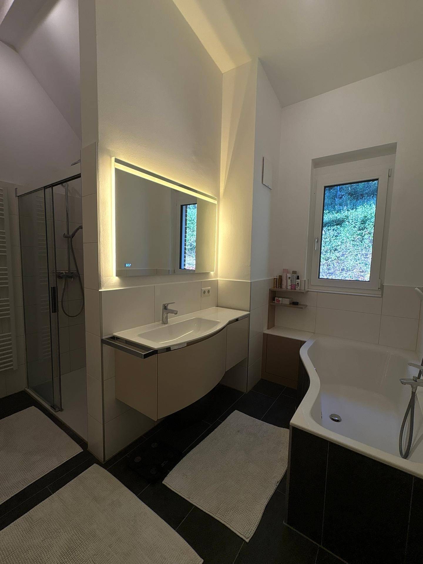 Modernes Badezimmer mit Badewanne, Dusche, beleuchtetem Spiegel und dunklen Fliesen.