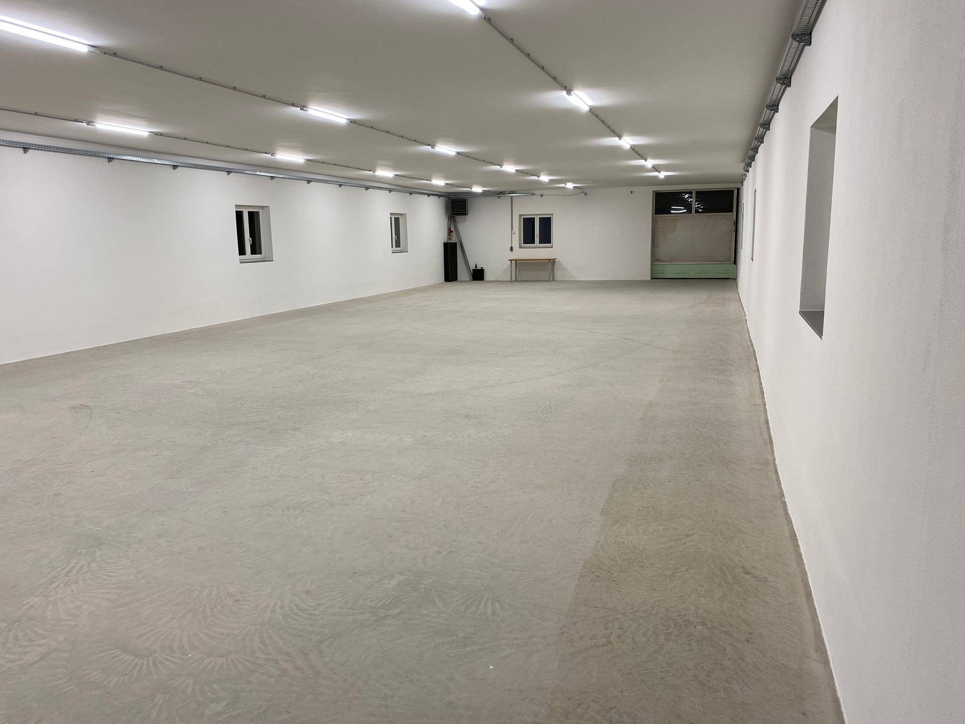 Geräumiger, leerer Lagerraum mit Betonboden, weißen Wänden und einer kleinen Arbeitsfläche im Hintergrund, gut beleuchtet.