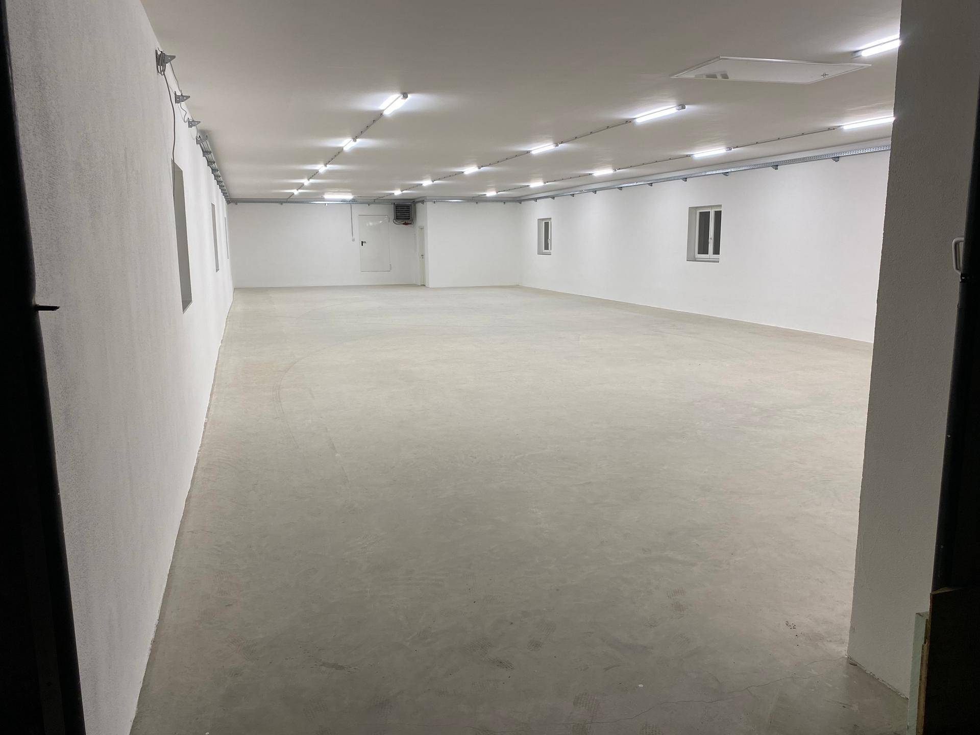 Langer, heller Raum mit glattem Betonboden und weißen Wänden, ausgestattet mit zahlreichen Deckenleuchten und kleinen Fenstern.