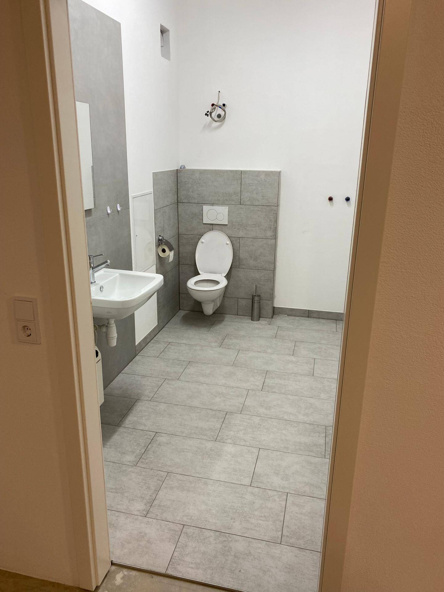 Modernes WC mit grauen Fliesen an Boden und Wänden, wandhängendem WC und einem Waschbecken mit Unterschrank.