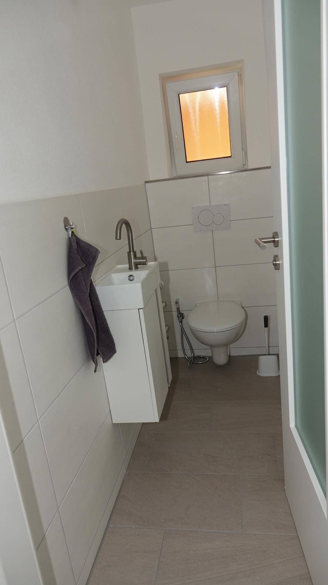 Separates Gäste-WC mit wandhängender Toilette, kleinem Waschbecken und Fenster für Belüftung.