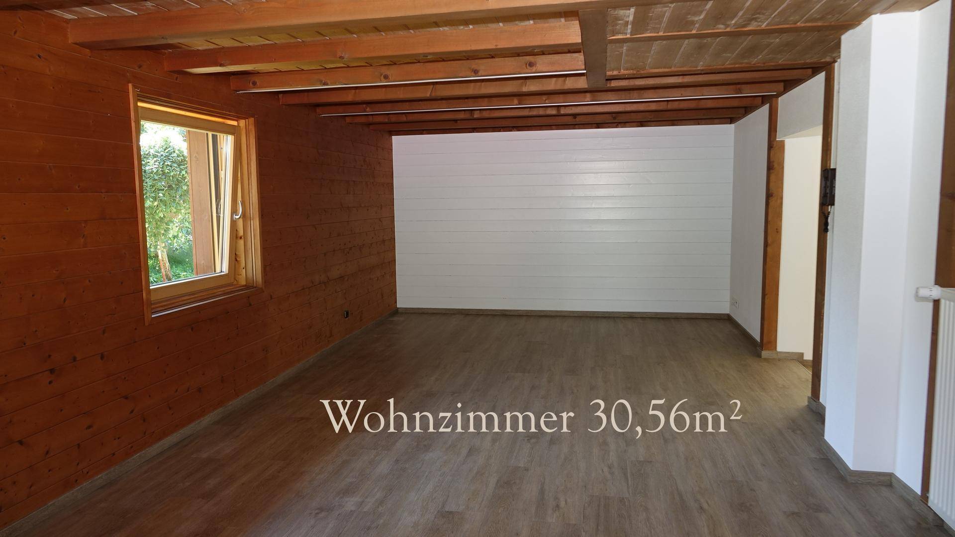 Geräumiges Wohnzimmer mit Holzwänden, Holzbalkendecke und Laminatboden, teilweise weiß gestrichen.