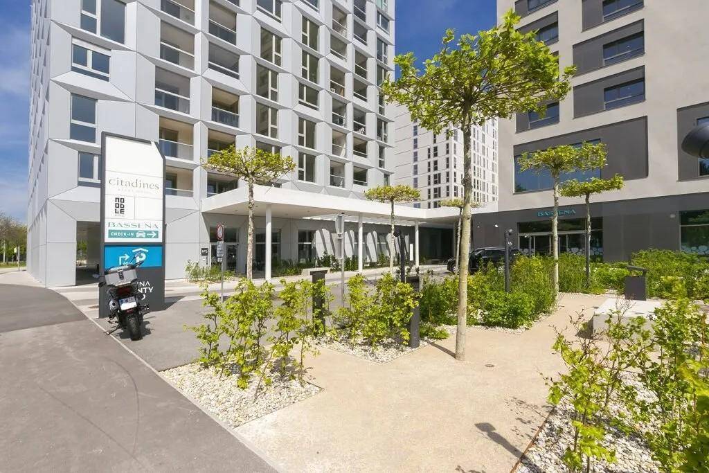 Eingangsbereich eines modernen Gebäudekomplexes mit gepflegter Außenanlage und kleinen Bäumen.