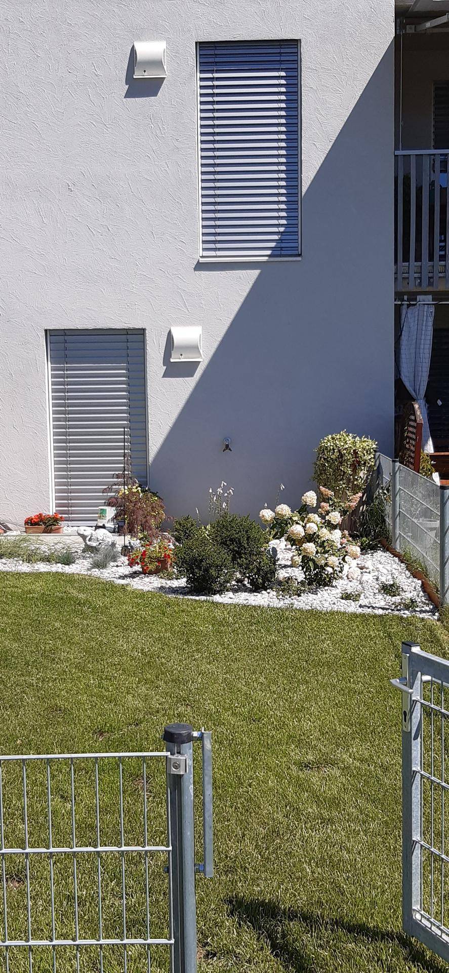 Gebäudefassade mit Fenster und Tür im Erdgeschoss, umgeben von einem gepflegten Gartenbeet mit weißen Steinen.