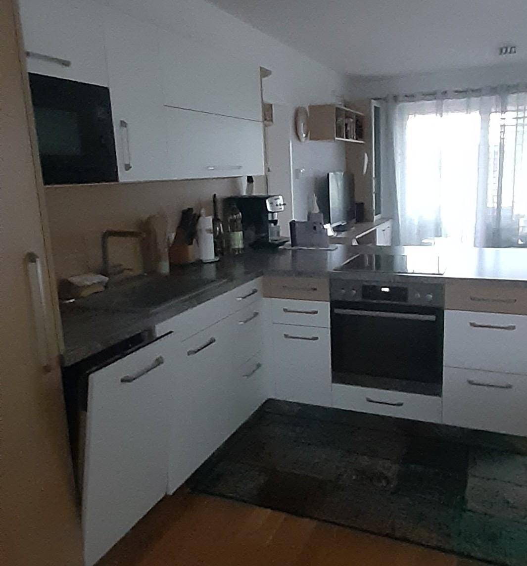 Küchenzeile mit integriertem Backofen, weißen Schränken und Blick auf ein Fenster mit Vorhängen im Hintergrund.