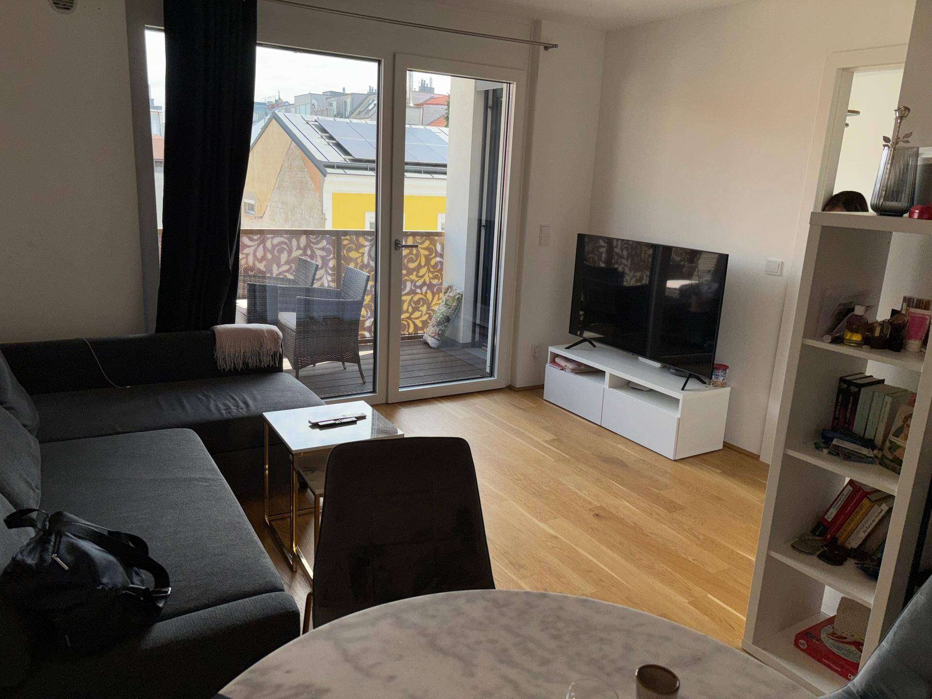 Helles Wohnzimmer mit Sofa, Fernseher und Zugang zu einem Balkon mit Blick auf die Umgebung.
