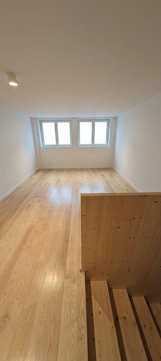Heller Raum im Dachgeschoss mit Holztreppe, schrägen Wänden und hohen Fenstern.