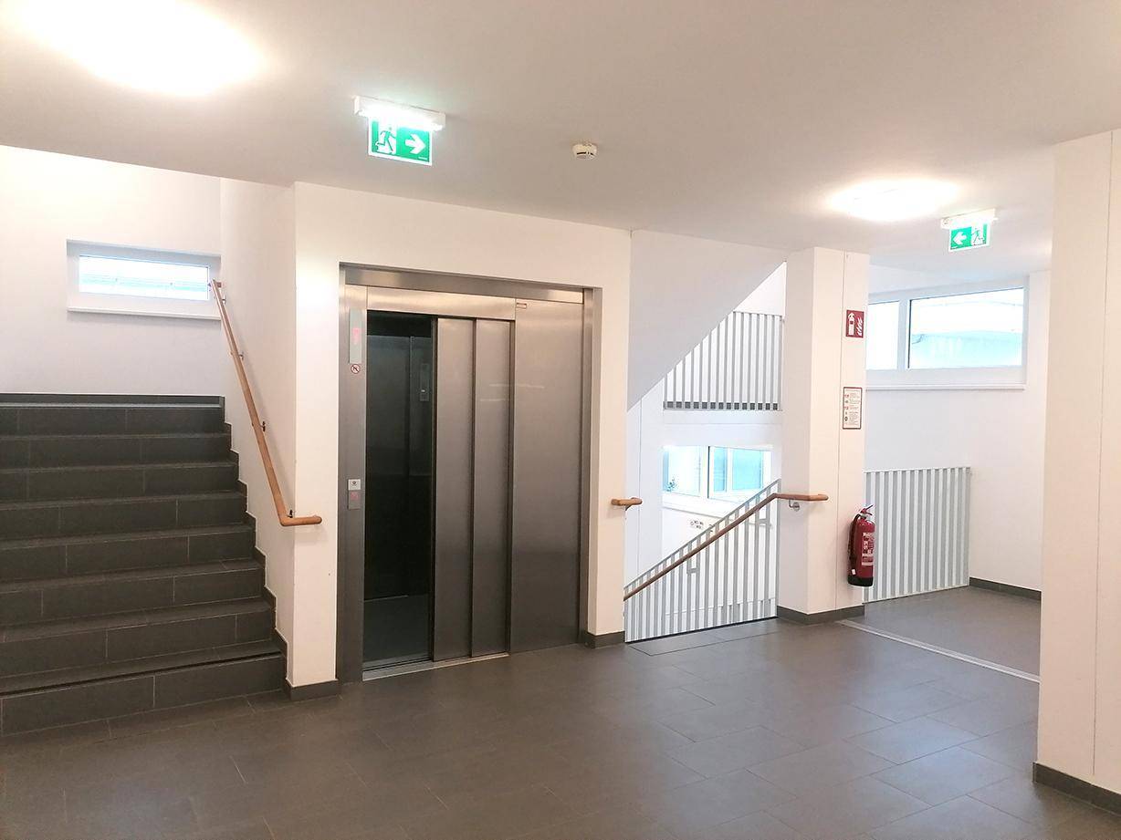 Heller Treppenaufgang mit Aufzug und Fliesenboden im modernen Gebäude.