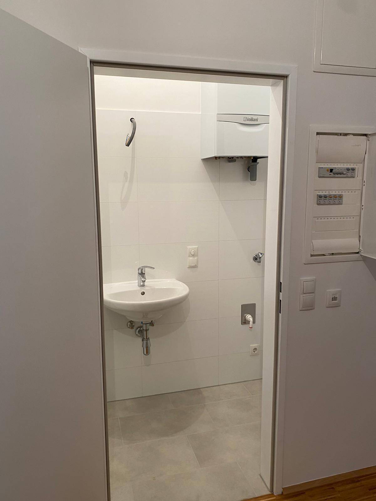 Kleiner Toilettenraum mit Waschbecken, weißen Fliesen und einem Warmwasserbereiter an der Wand.