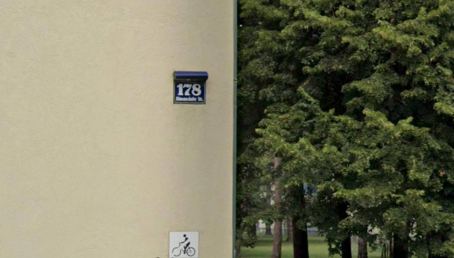 Nahaufnahme der Hausnummer 178 an einer hellen Gebäudewand neben einem großen Baum.