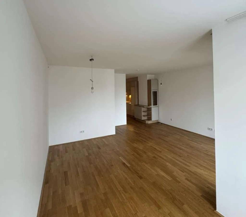Geräumiger Wohnbereich mit hellem Parkettboden und offenem Übergang zur Küche im Hintergrund.