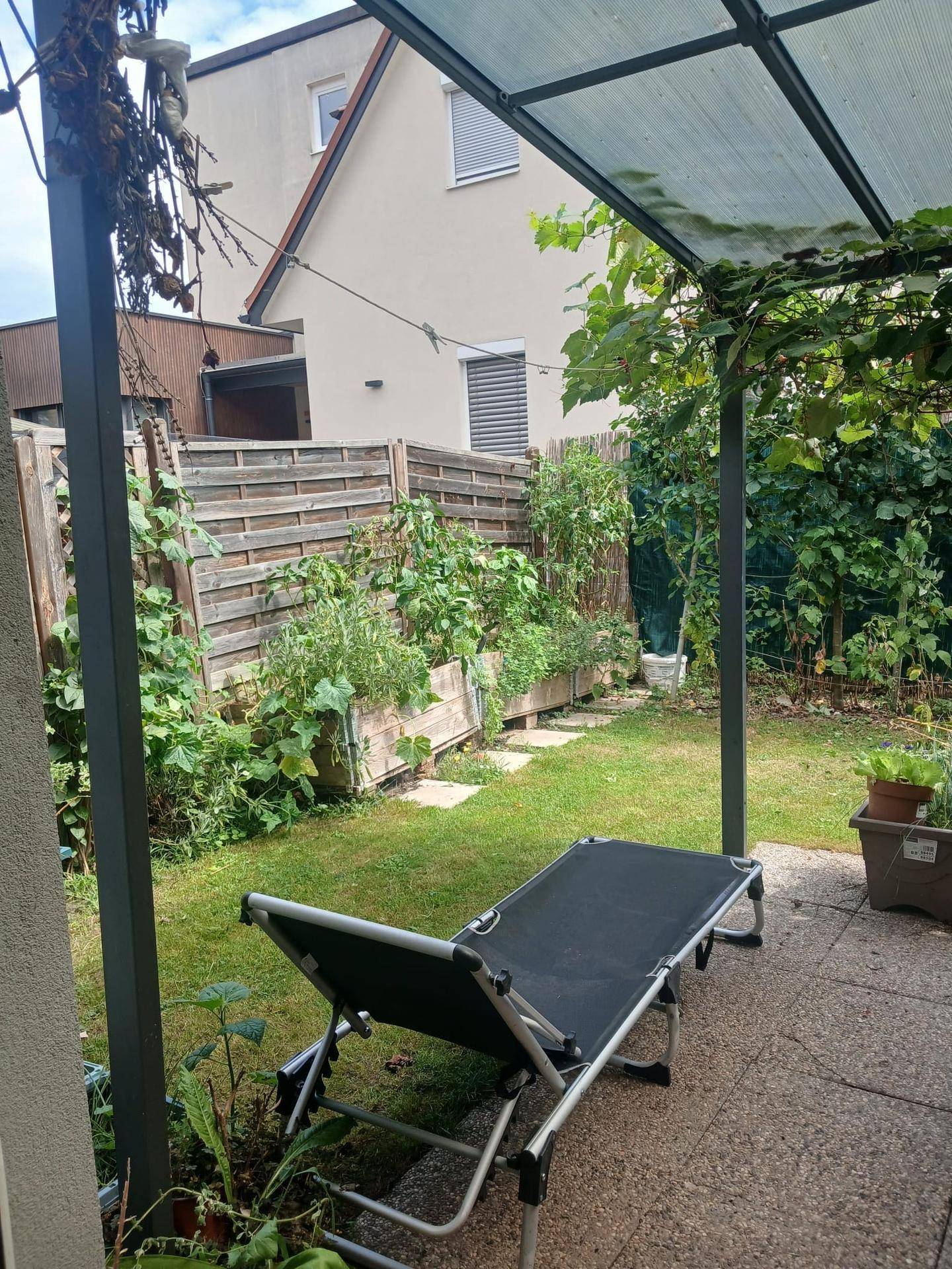Hinterhof mit Rasenfläche, Hochbeeten und einer überdachten Terrasse mit Liegestuhl.