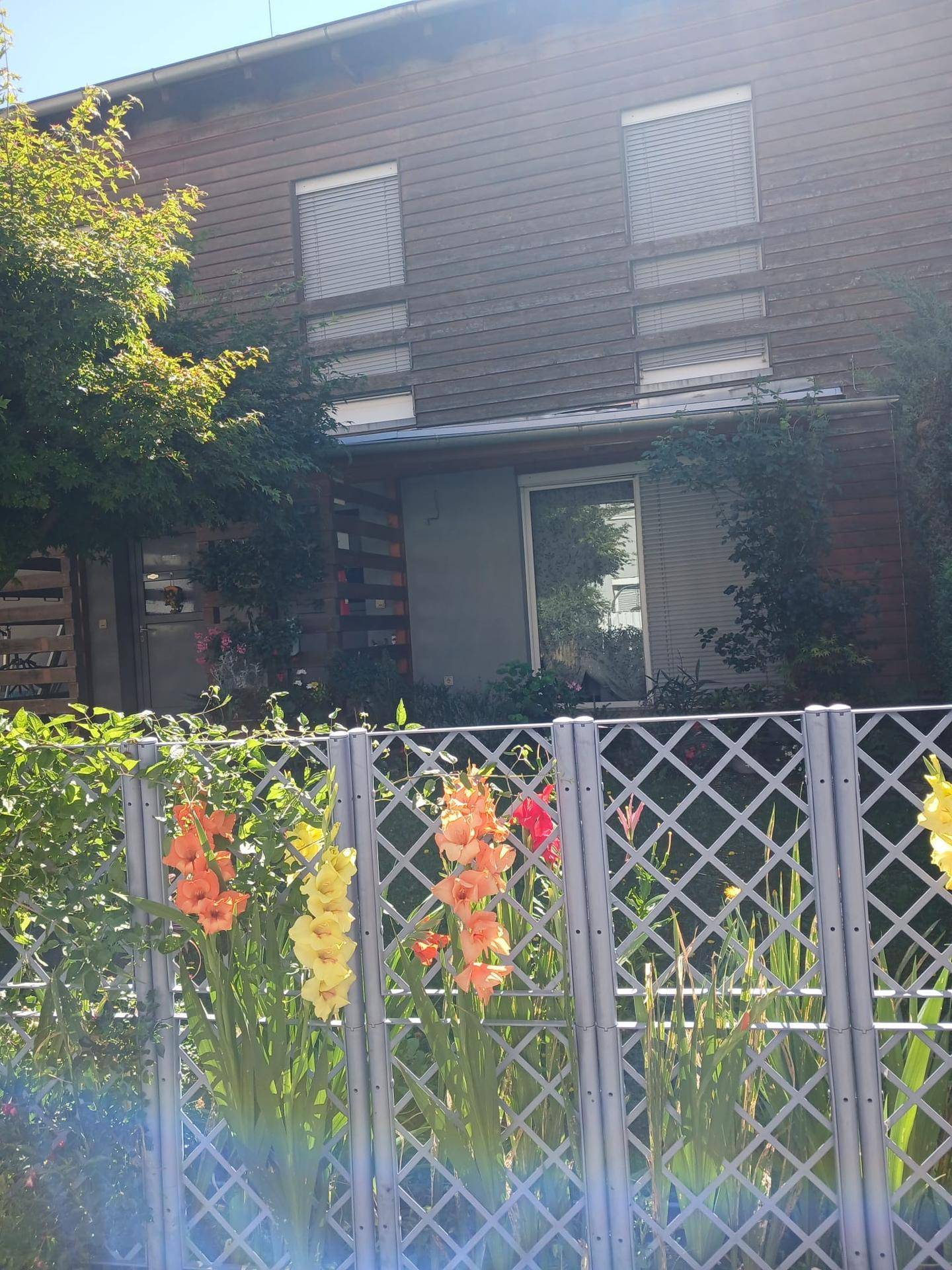 Hausfassade mit mehreren Fenstern und Rollläden, im Vordergrund ein Zaun mit bunten Blumen.
