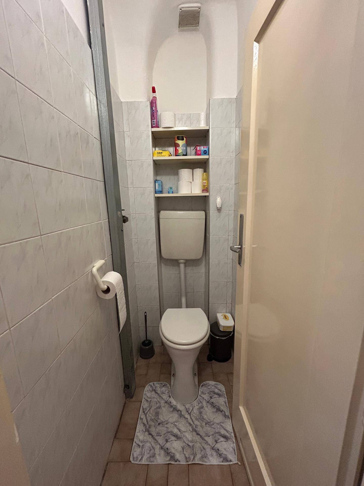 Separates WC mit Toilette, Fliesen an den Wänden und einem kleinen Regal für Utensilien.