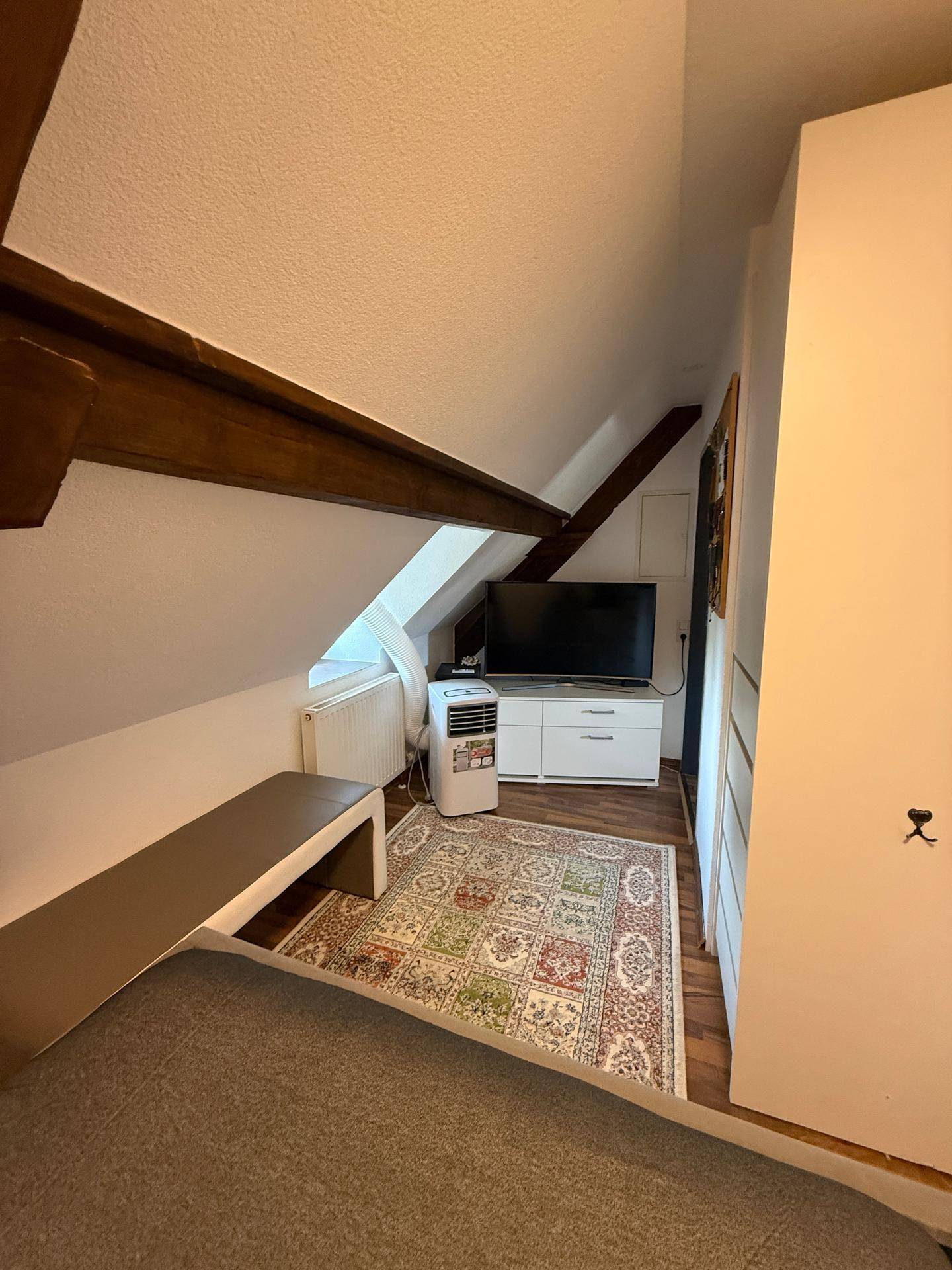 Gemütliches Dachzimmer mit schräger Decke, Fernseher, Sitzbank und gemustertem Teppichboden.