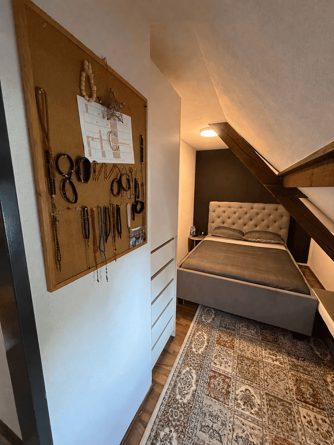 Schlafzimmer im Dachgeschoss mit schräger Decke, gepolstertem Bett und gemustertem Teppich.
