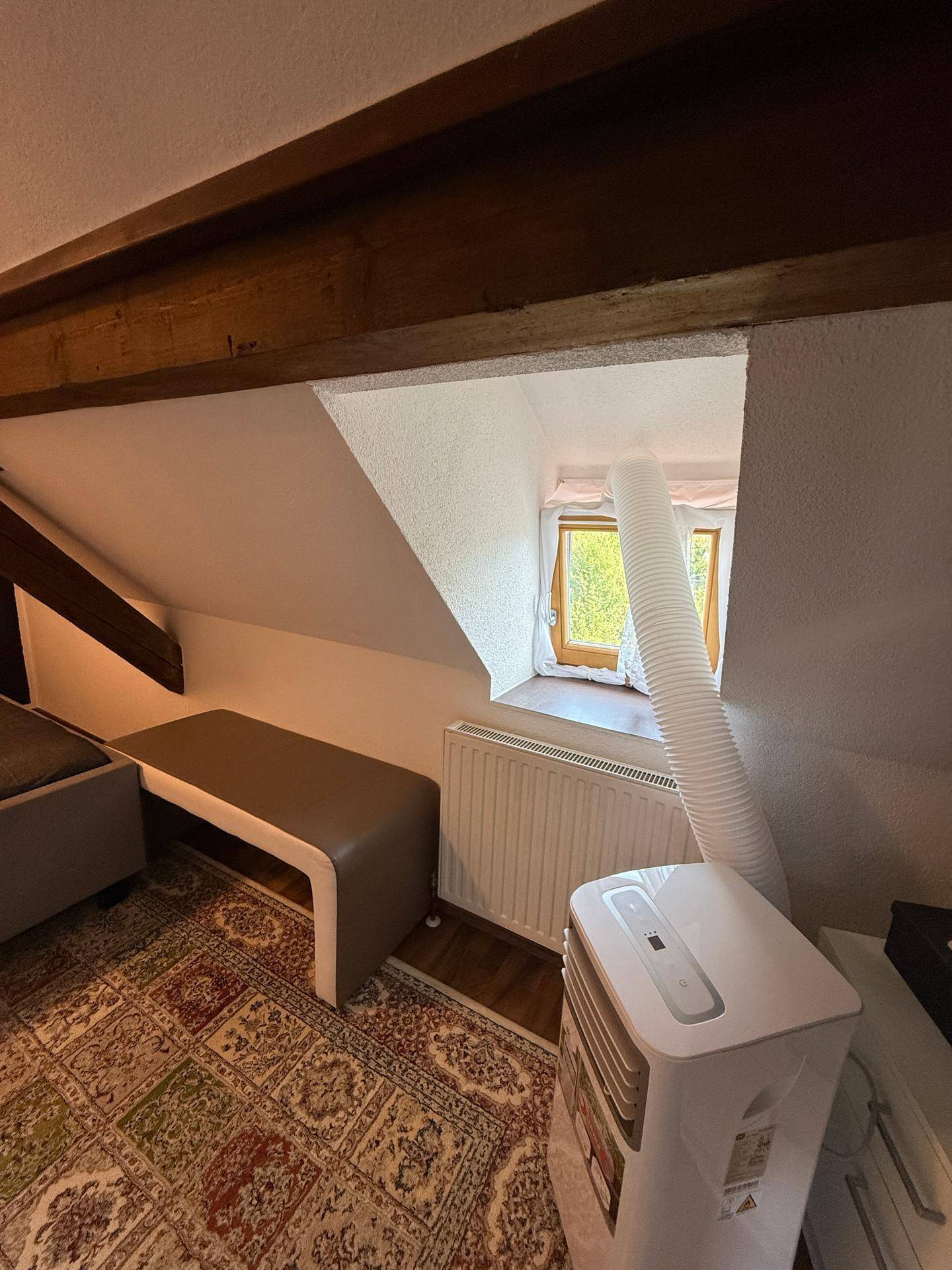 Dachzimmer mit schräger Decke, Fenster, Heizkörper und mobiler Klimaanlage, unter Holzbalken.