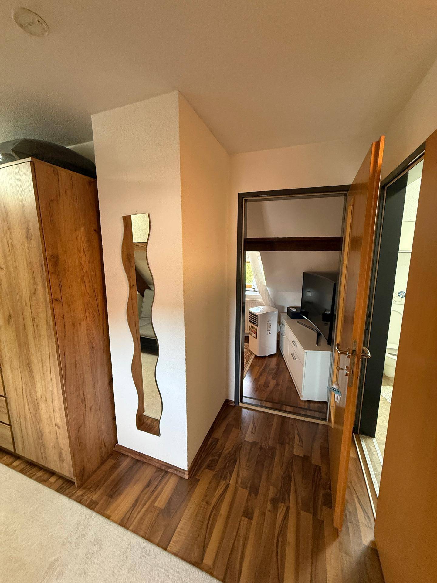 Flur mit Holzschrank und Spiegel, der zu weiteren Zimmern führt, darunter ein Raum mit Fernseher.