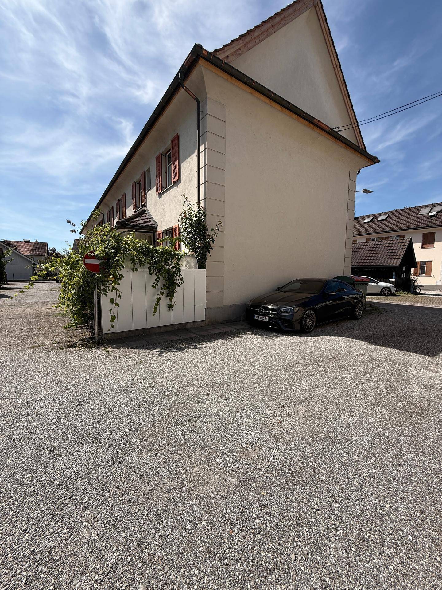 Seitenansicht des Hauses mit Giebeldach, roten Fensterläden und einem geparkten Auto auf dem Schotterweg.