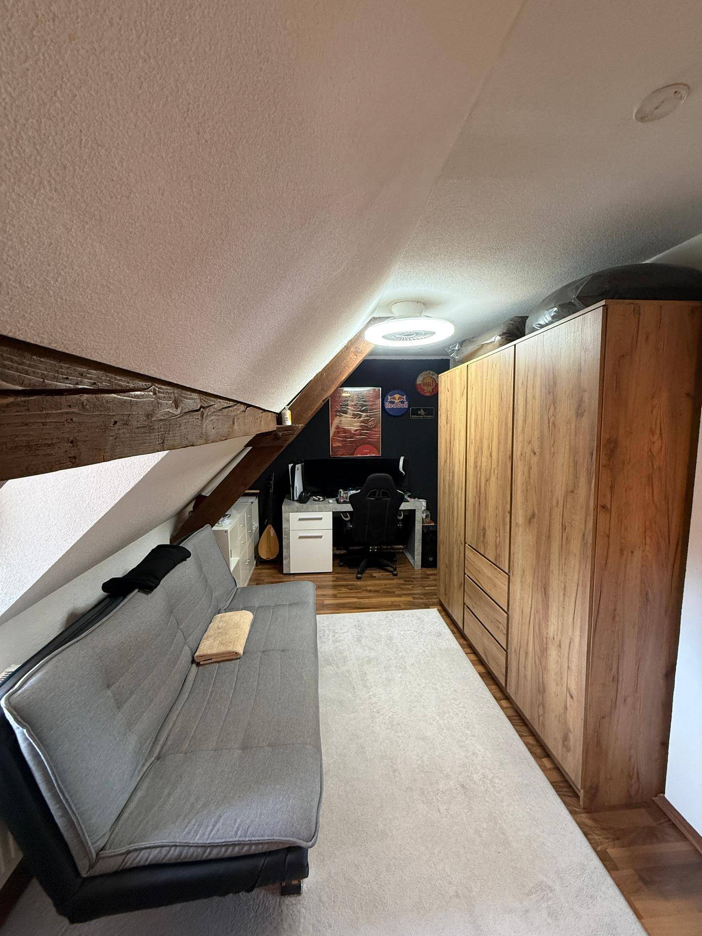 Gemütliches Dachzimmer mit schräger Decke, Sofa, Arbeitsbereich und großem Holzschrank.