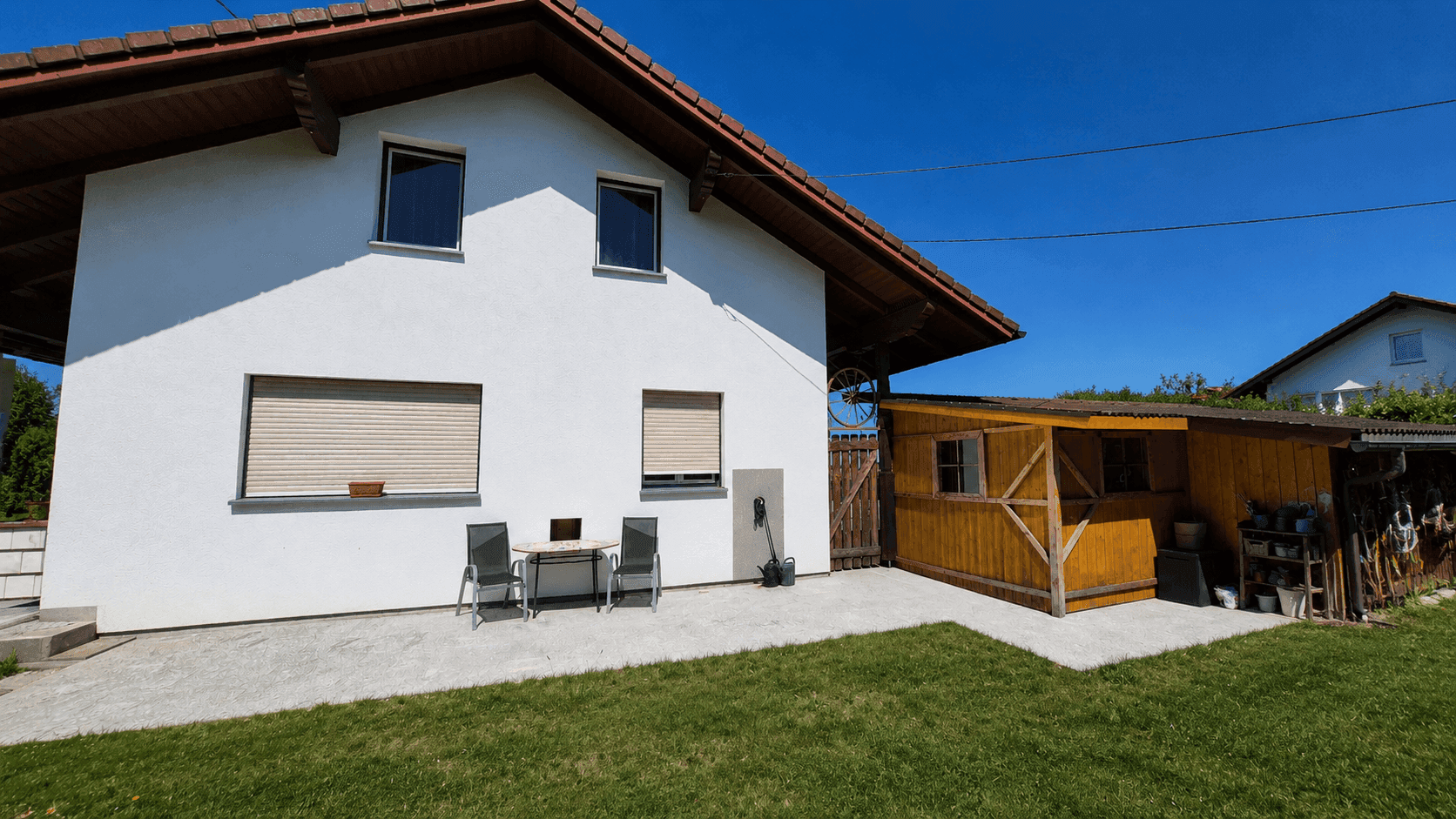 Rückseite des Hauses mit kleiner Terrasse, Garten und Holzschuppen unter blauem Himmel.