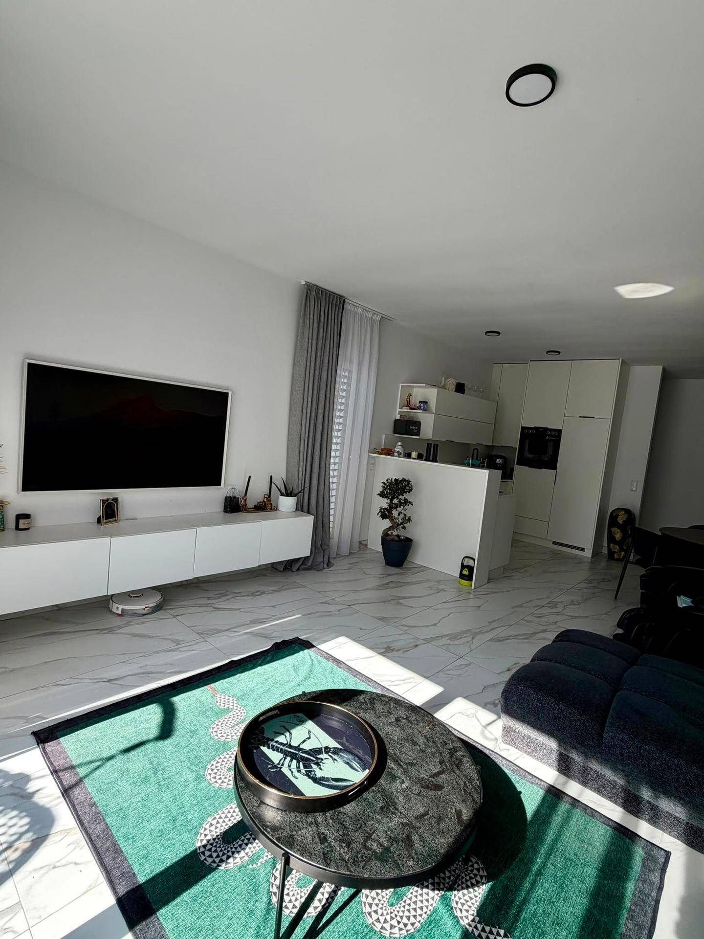 Helles Wohnzimmer mit großem Fernseher, modernem Teppich und Blick zur offenen Küche.