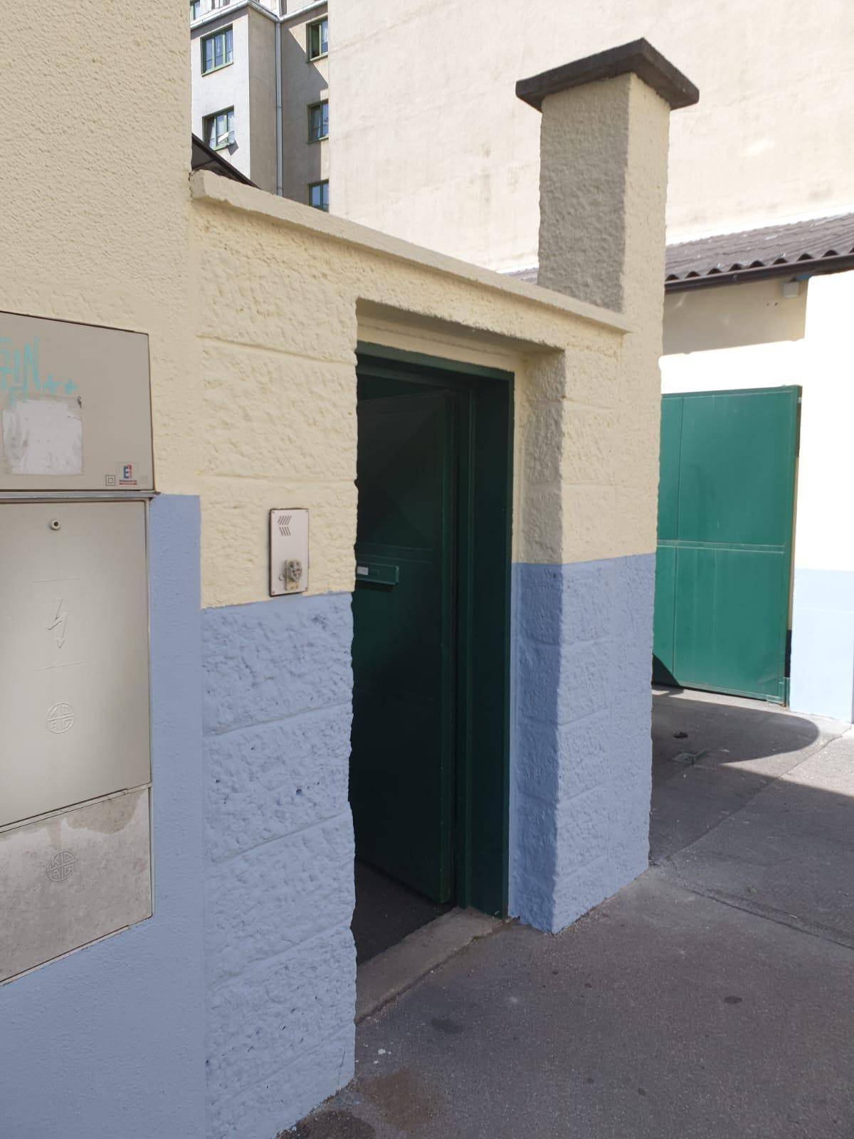 Eingangsbereich eines Gebäudes mit beiger und blauer Fassade, einer grünen Tür und einem kleinen Schornstein.