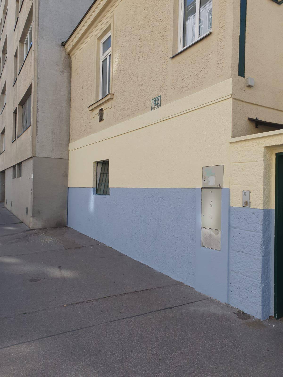 Fassade eines Gebäudes mit beiger und blauer Farbgebung, kleinen Fenstern und einem Gehweg davor.