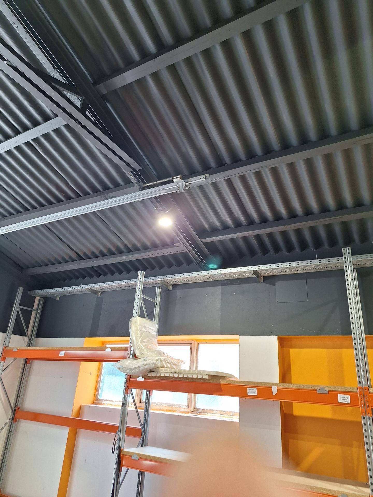 Blick auf die Deckenkonstruktion eines Lagerraums mit Metallträgern und Wellblechdach sowie Regalsystemen.