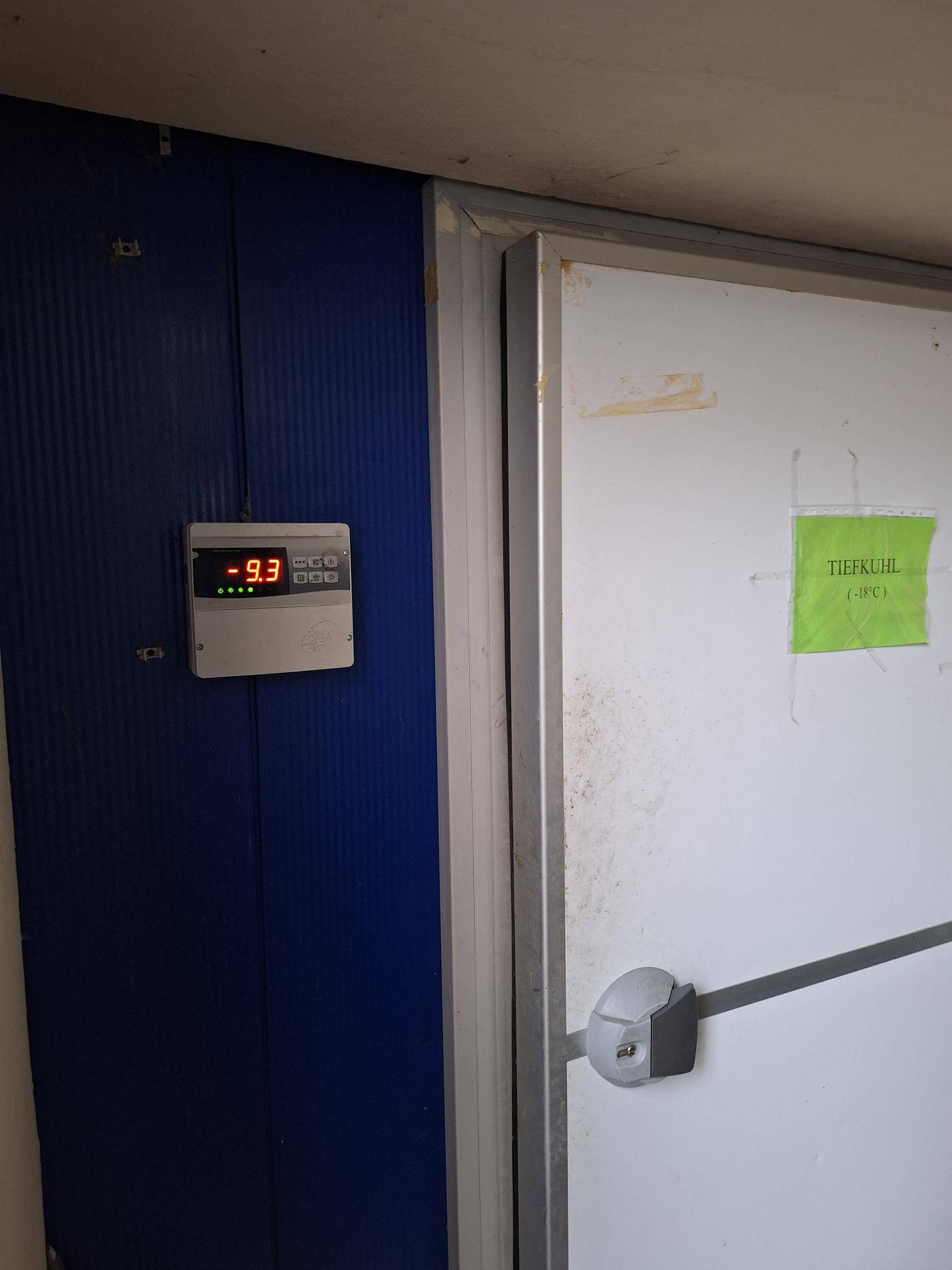 Digitalanzeige mit -9,3 Grad Celsius neben einer weißen Tiefkühltür in einem Lagerbereich.