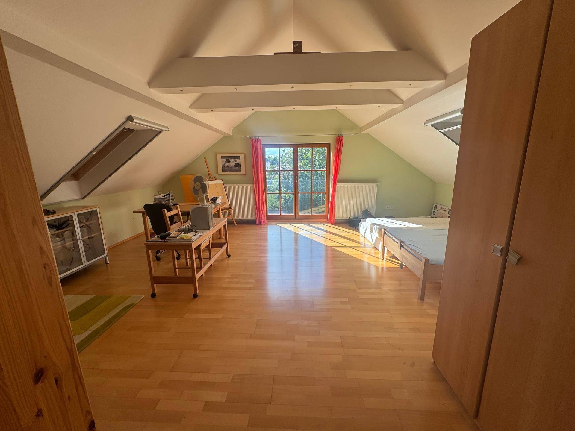Helles Dachzimmer mit Holzboden, Dachschrägen und Zugang zu einem Balkon oder einer Terrasse.