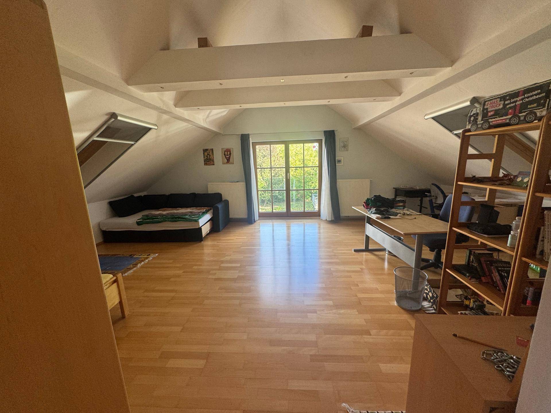 Geräumiges Dachzimmer mit Schlafsofa, Arbeitsbereich, Holzboden und viel Tageslicht durch große Fenster.