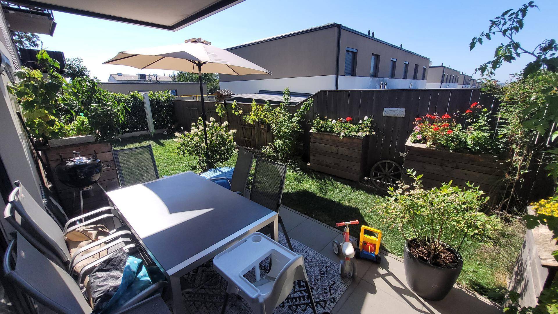 Sonniger Garten mit Terrasse, Esstisch und Sonnenschirm, umgeben von grünen Pflanzen und einem Zaun.