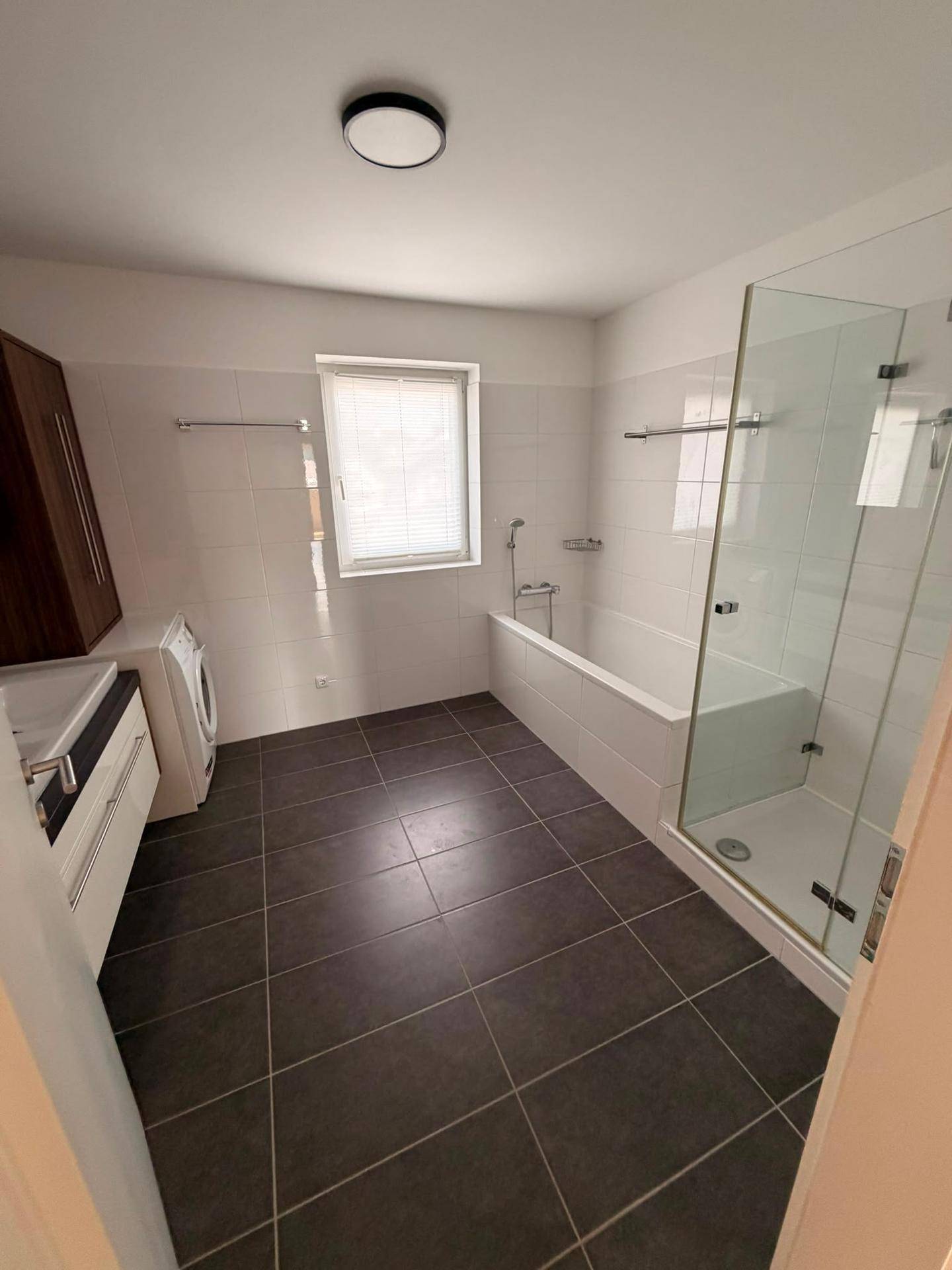 Helles Badezimmer mit Fenster, Badewanne, Duschkabine und Anschluss für Waschmaschine.