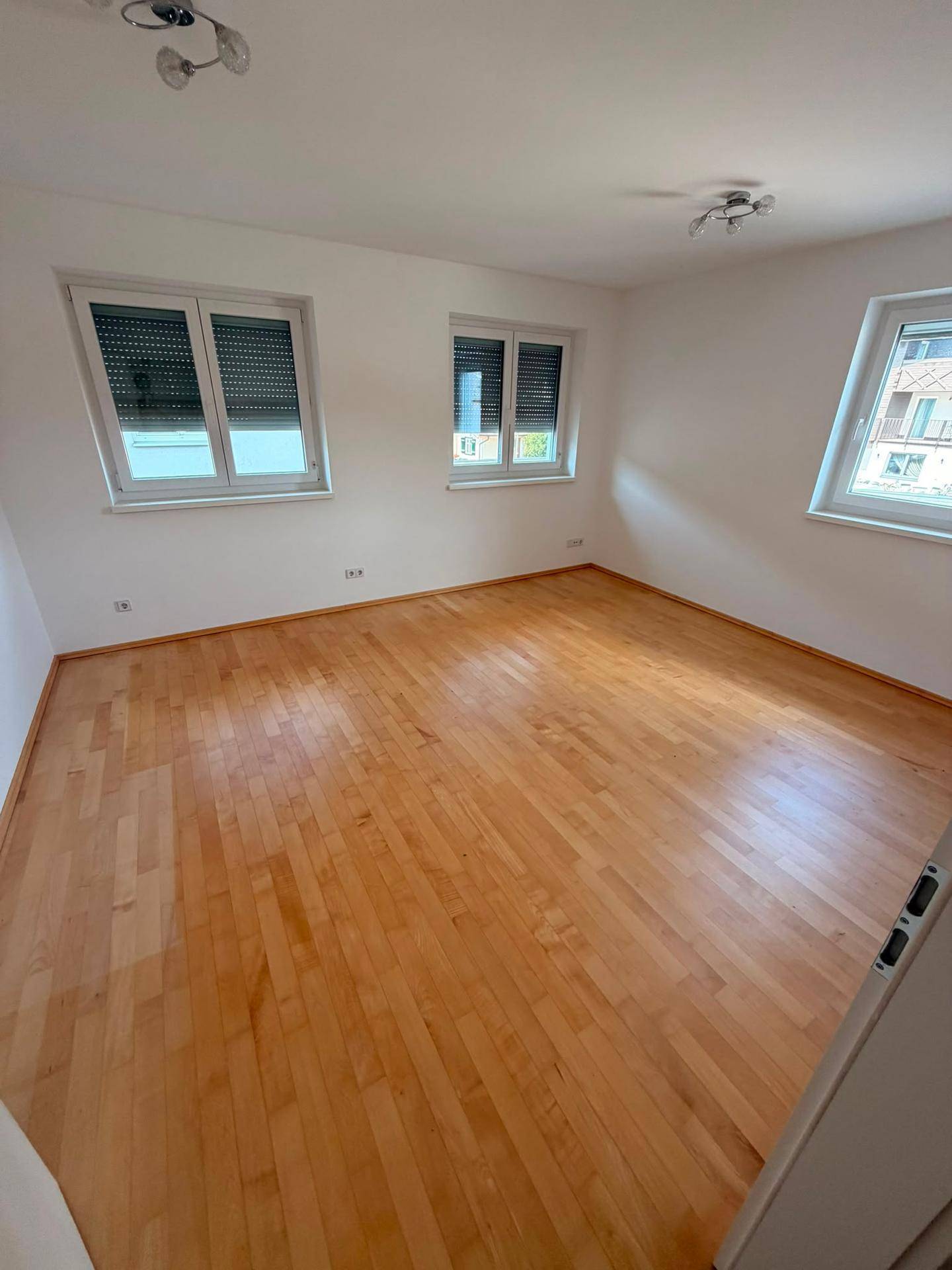 Helles, leeres Zimmer mit Holzboden und drei Fenstern, die viel Tageslicht bieten.