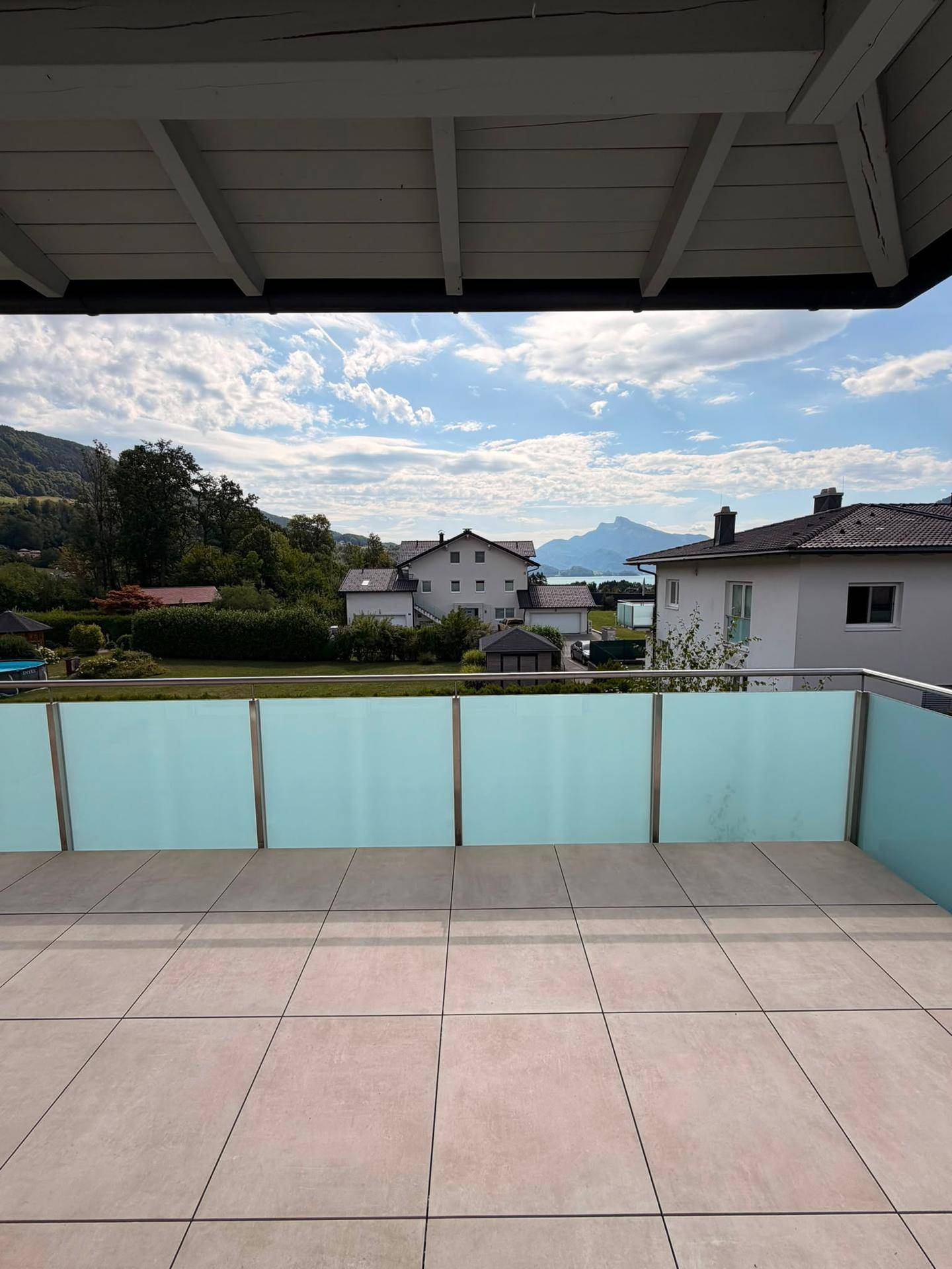 Geräumige Terrasse mit Fliesenboden und Glasgeländer, bietet weiten Blick auf See und Berge.