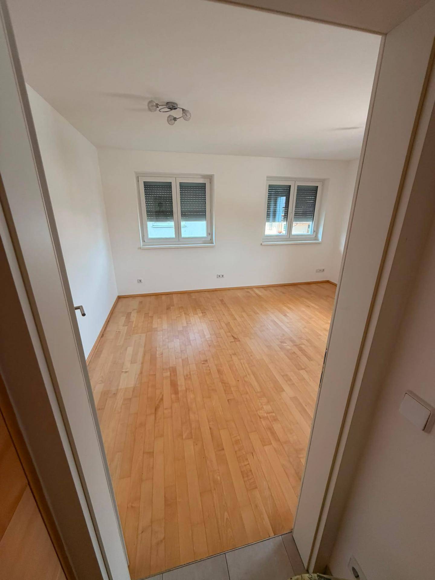 Leeres Zimmer mit hellem Holzboden und zwei Fenstern, ausgestattet mit Rollläden.