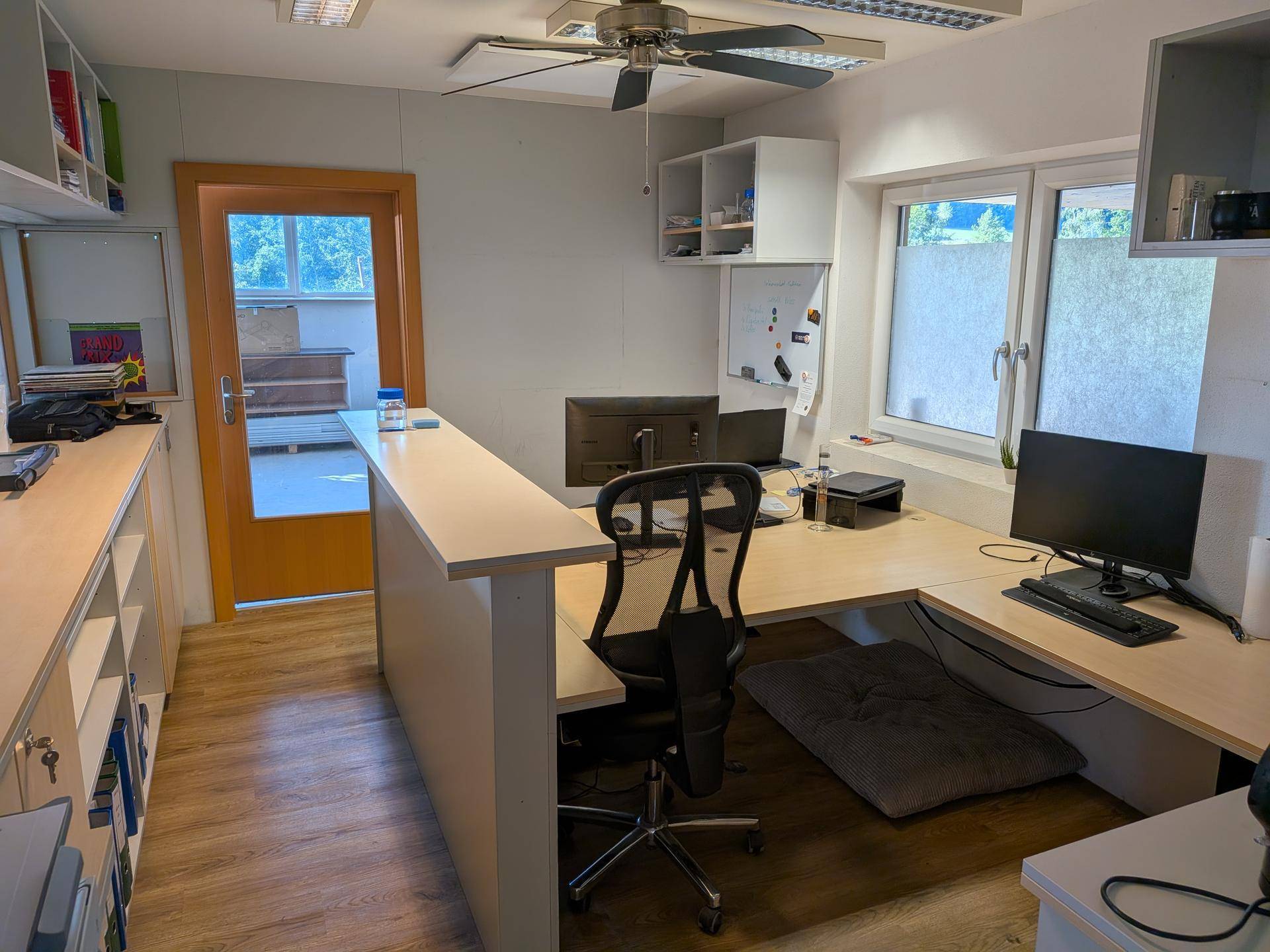 Modernes Büro mit großem L-förmigem Schreibtisch, Deckenventilator und Regalen, ideal für konzentriertes Arbeiten.
