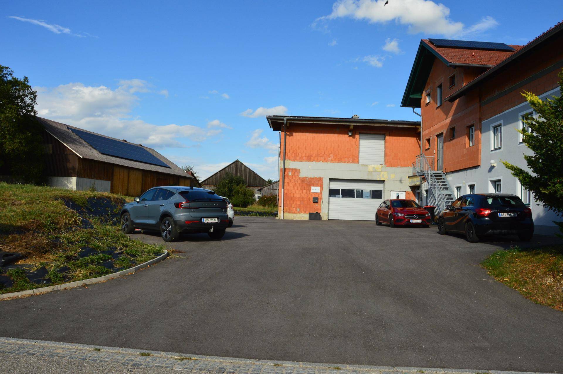 Außenansicht eines Gebäudekomplexes mit asphaltierter Zufahrt und geparkten Fahrzeugen, umgeben von Grünflächen.