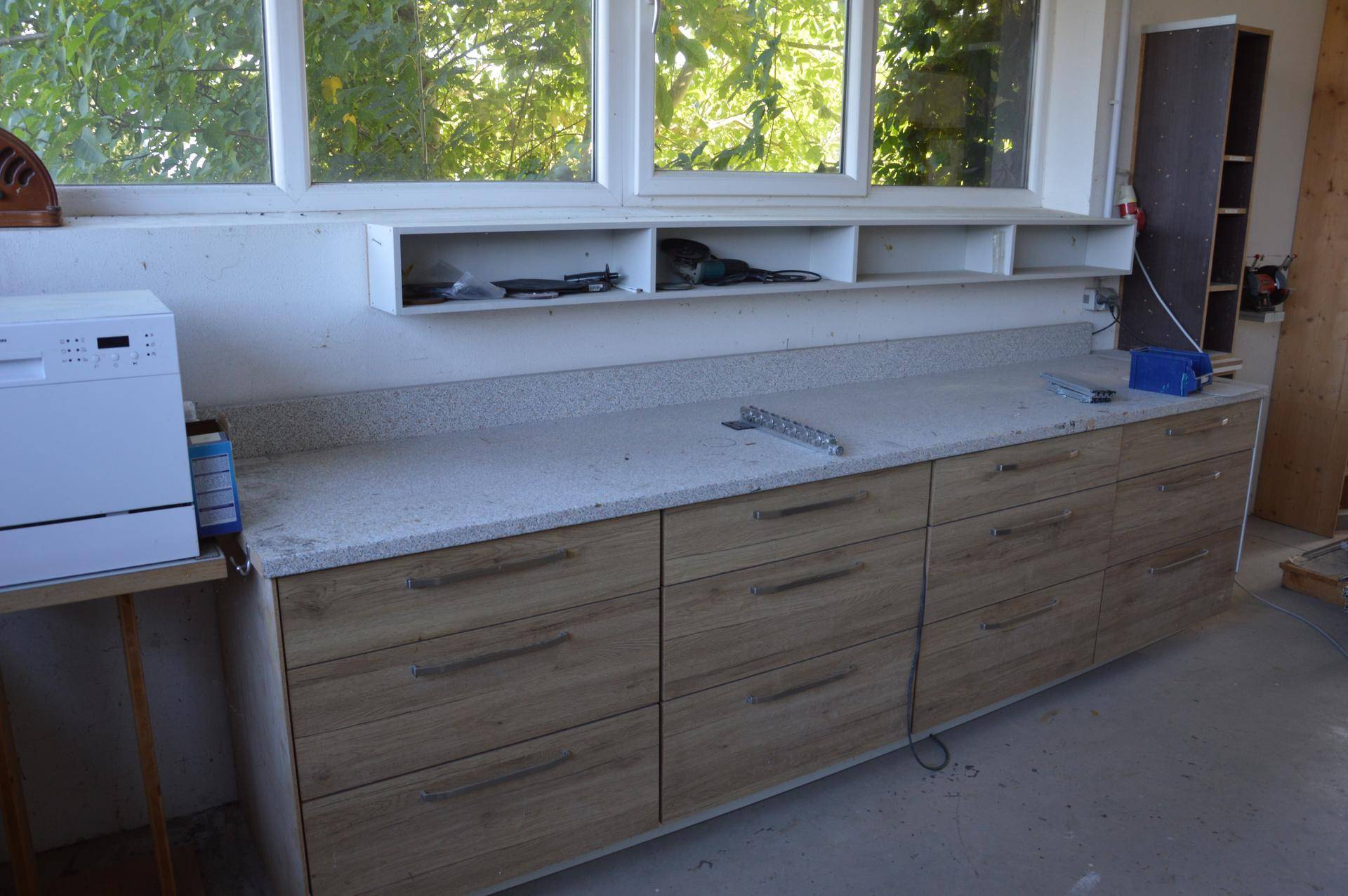 Langer Arbeitstisch mit Holzschubladen und heller Arbeitsplatte unter Fenstern, inklusive kleinem Geschirrspüler.