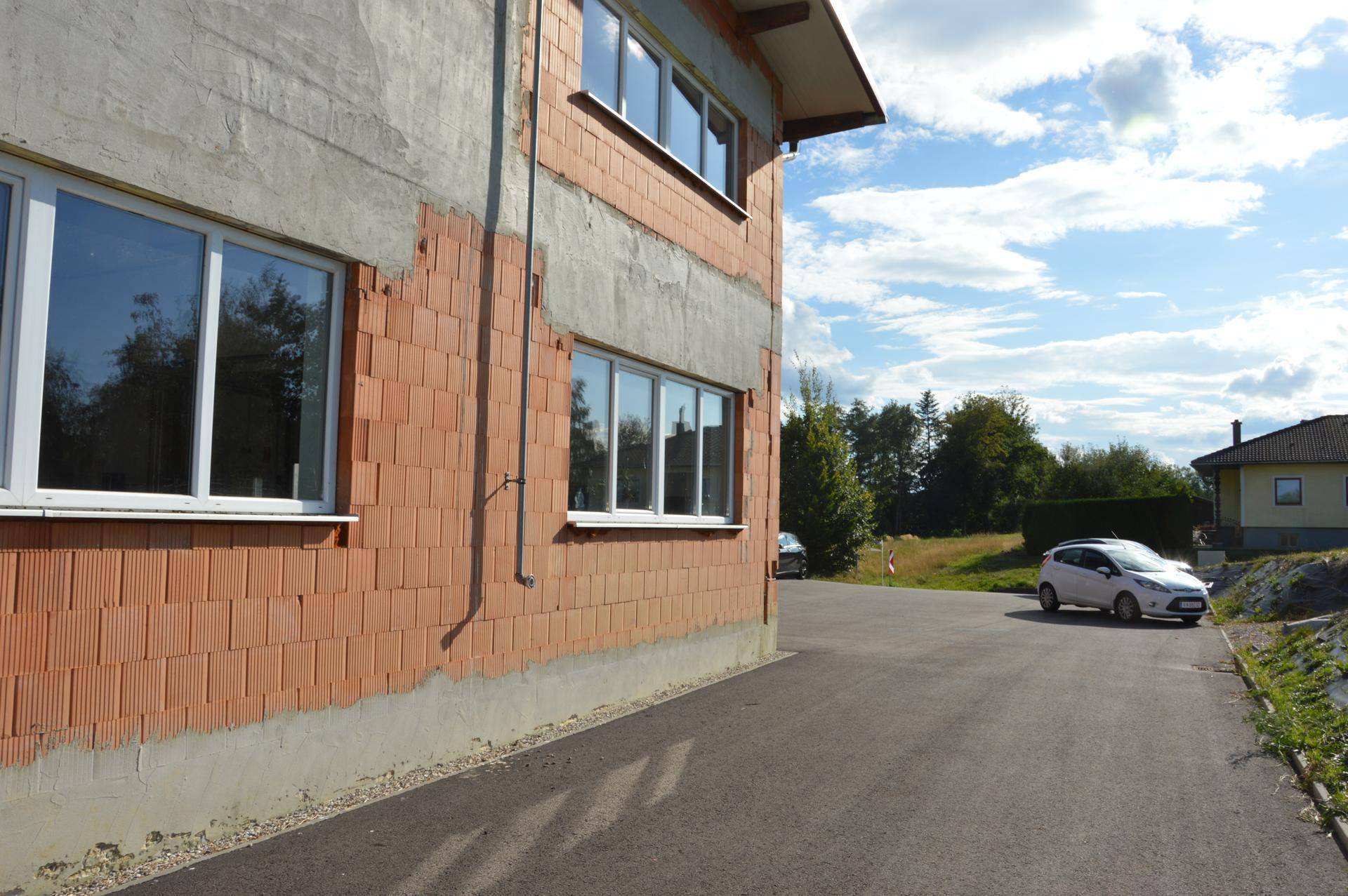 Seitenansicht eines Gebäudes im Bau mit Ziegelwänden und neuen Fenstern, umgeben von Natur.